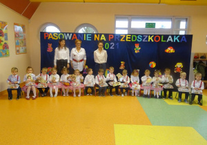 Grupa dzieci siedzi na krzesełkach, na kolanach trzymają upominki. Za dziećmi stoi pani dyrektor Maria Królikowska, pani Kinga i pani Ewelina.